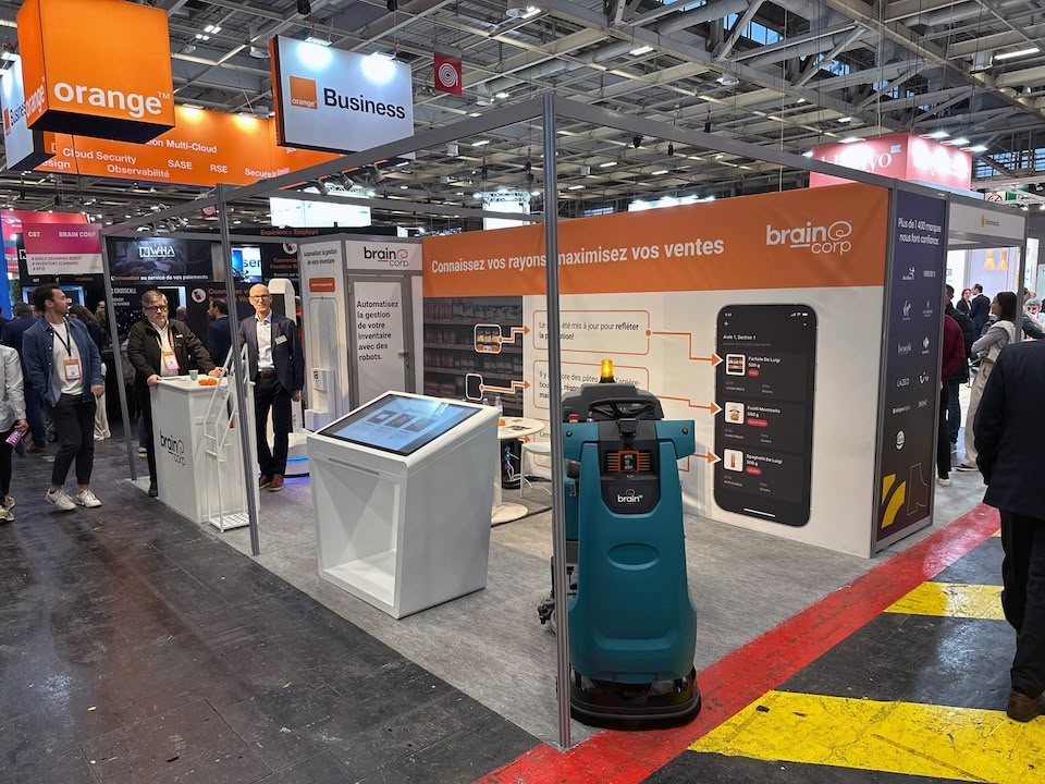 A BrainCorp trade show booth with orange "Connaissez vos rayons" panel, kiosk and floor-cleaning robot